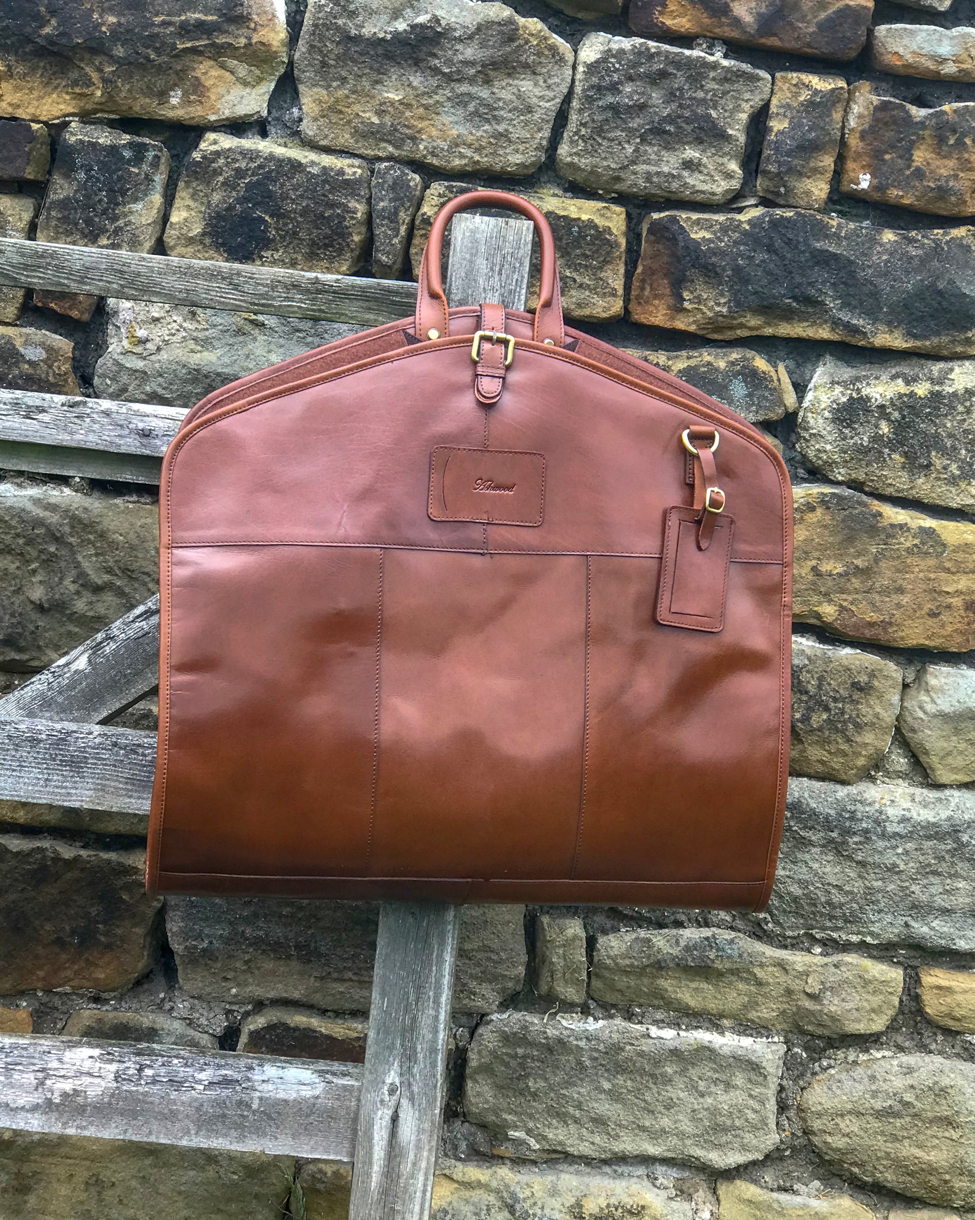 Ashwood chestnut leather Suit Carrier hung up open against stone wall
